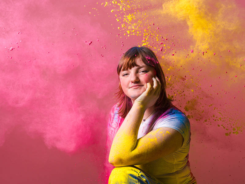 A College age student crouched on the floor on a pink background. She is covered in brightly coloured powder in pink and yellow.