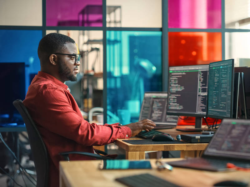African American Man Writing Lines Of Code On Desktop Computer With Multiple Monitors and Laptop in Creative Office
