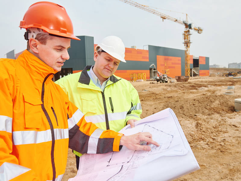 Two men in hard hats and hi-vis jackets looking at plans on a construction site.