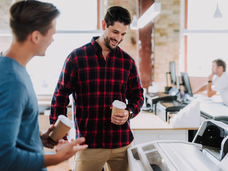 Cheerful man drinking coffee while standing with his colleague near a printer.